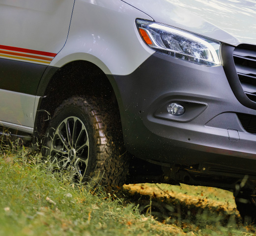 A Mercedes-Benz Camper Van parked on a steep hill with a person sitting next to it fixing their bike. 